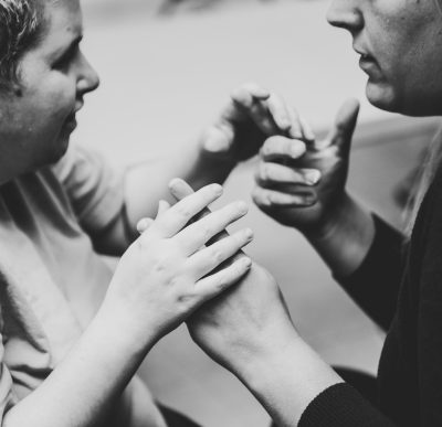 A black and white photo of the side profiles of 2 people using ASL hand over hand. The person on the left has their hands on top of the hands of the person on the right. The person on the left shows the side of their face and upper body the person on the right has only from their nose down showing.