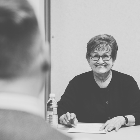 Black and white photo of a woman sitting at a table. She has short hair, glasses and is wearing a black sweater. There is a water bottle and paper in front of her. Across from her is a man facing her.
