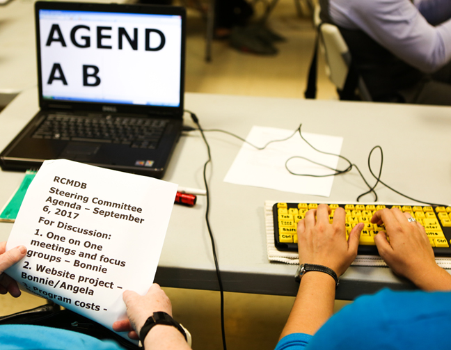 Coloured photo of a black laptop on top of a white table with a keyboard plugged in and a persons hands typing on the keyboard. The computer has big bold black letters. The person sitting in front of the computer is holding up an RCMDB Steering Committee September Agenda. Both people are wearing blue tops.