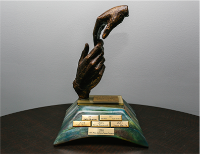 Coloured photo of the Evelyn Presley Award. 2 statue hands touching, one reaching from below and the other touching from above. The hands are on top of a beautifully hand made glass stand. The glass stand has an array of colours including green, blue, auburn and yellow. On the top of the stand is gold plated plaque, in front, there are 6 gold plated plaques and one gold plated plaque on the right side. On each of plaques are the names of the individuals who received the award.
